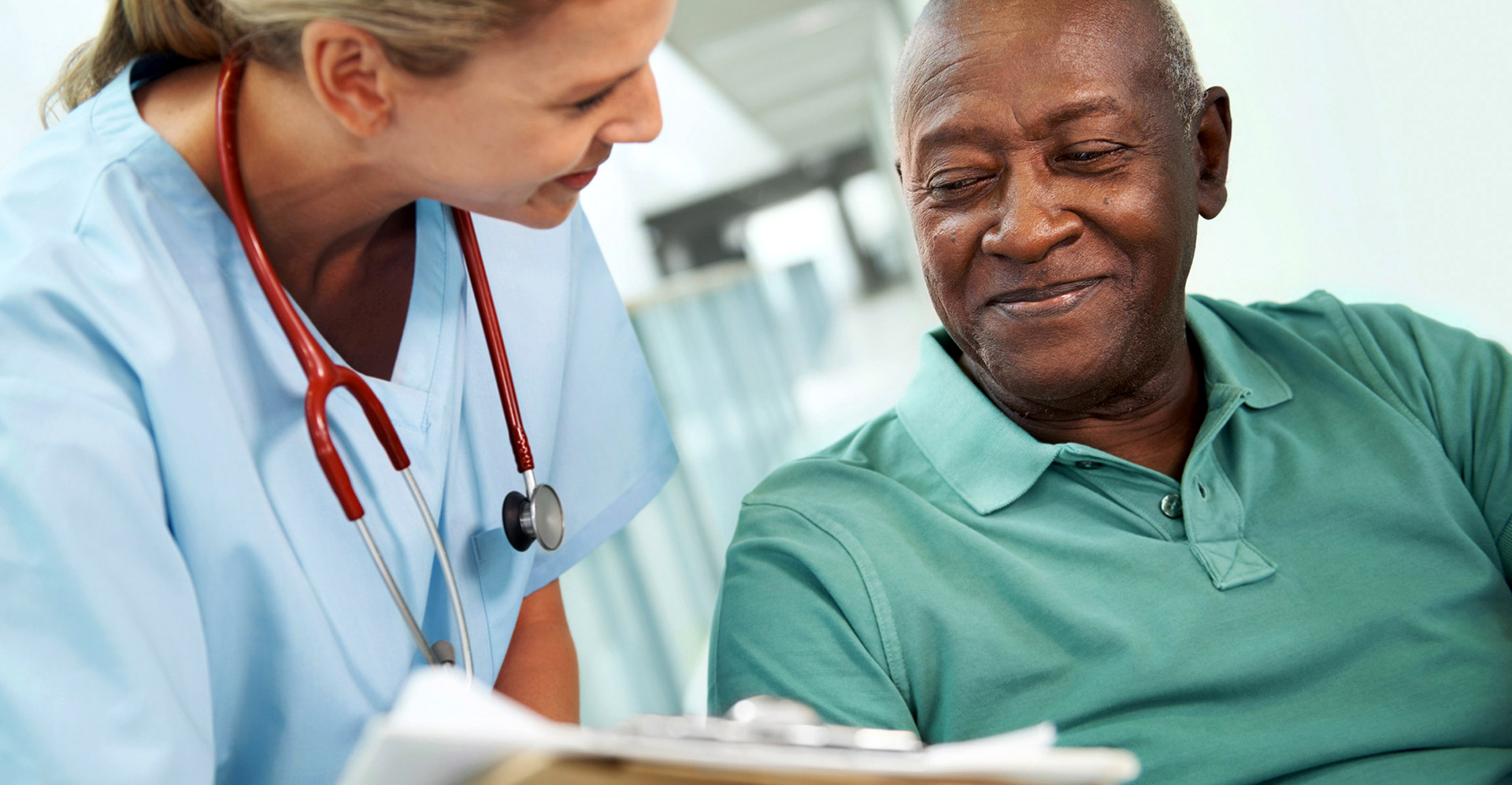 A healthcare working with a stethoscope around her neck bending down to discuss paperwork with a patient. The patient is elderly, smiling, and wearing a green polo shirt.