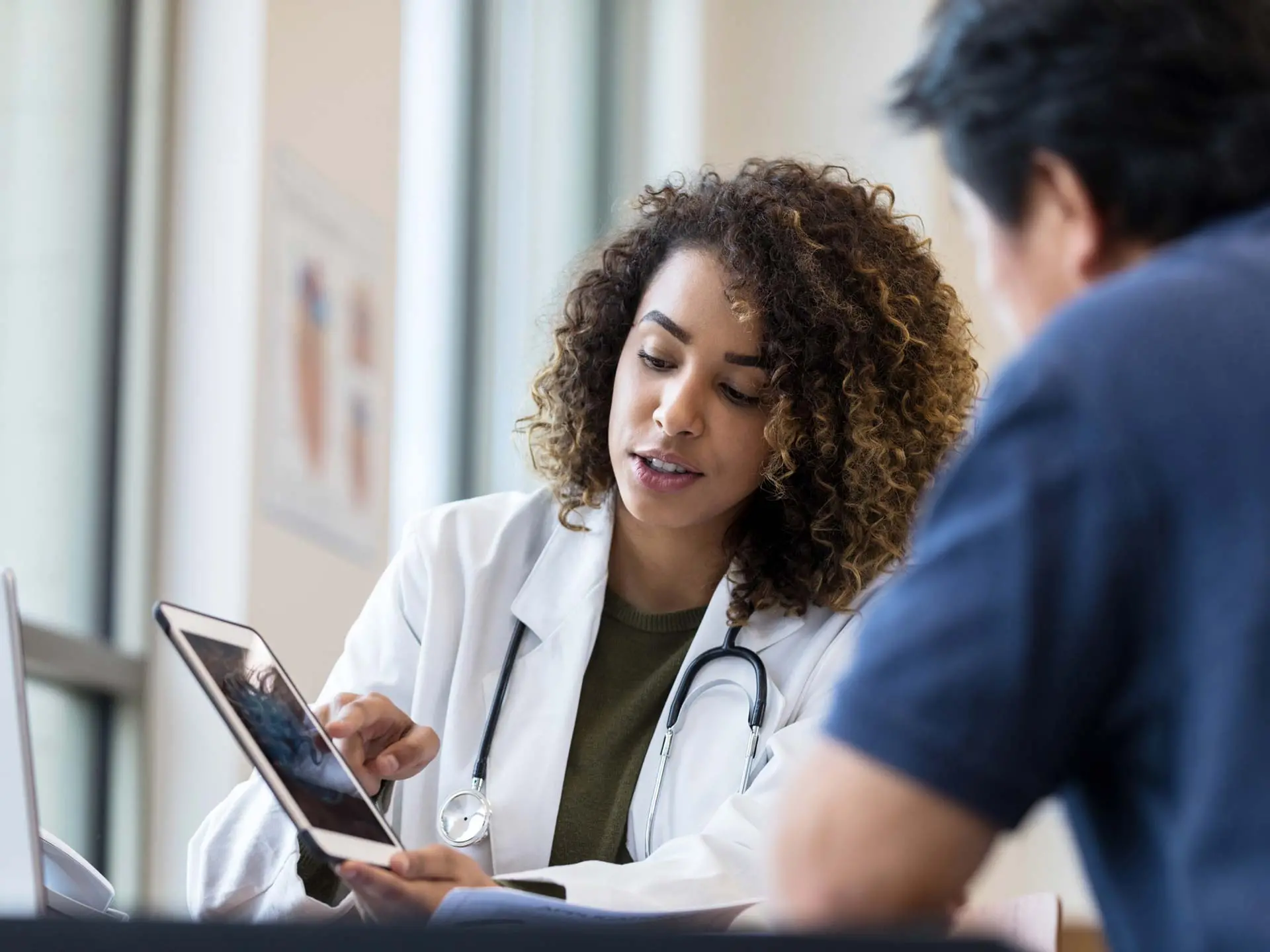 A healthcare professional working with a stethoscope around her neck bending down to discuss paperwork with a patient.