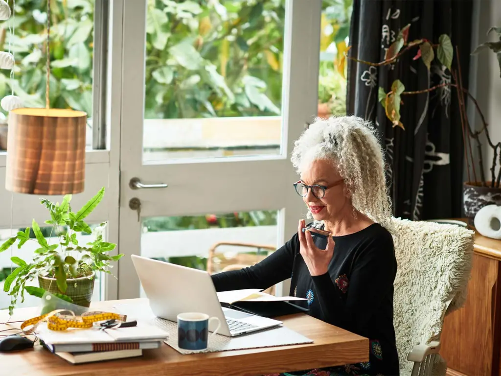Grey haired female entrepreneur multitasking at home, video conference, speaking and listening on mobile phone, wireless technology