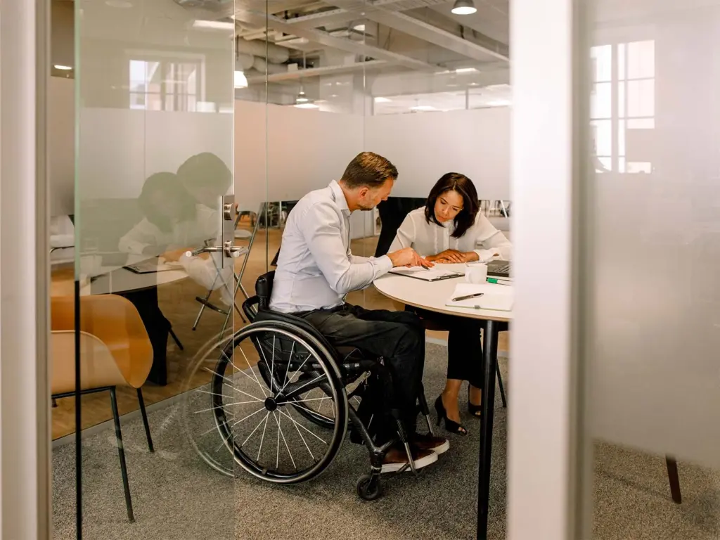 Employee in wheelchair and coworker look over data documents together in meeting room.