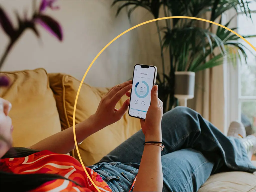 A relaxed woman lies back on a comfortable yellow sofa in a domestic environment. She holds a smart phone and uses a modern thermostat application to control the heating system within the house.