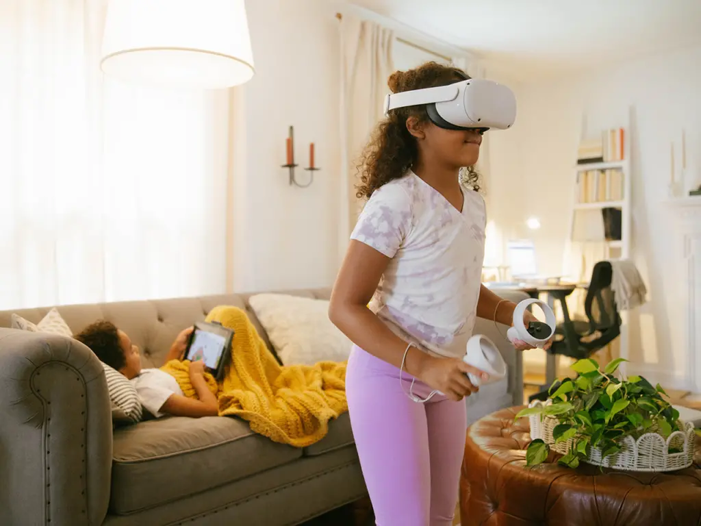 Little girl interacting with augmented reality and virtual reality headset while her brother is on the couch behind her with a tablet.