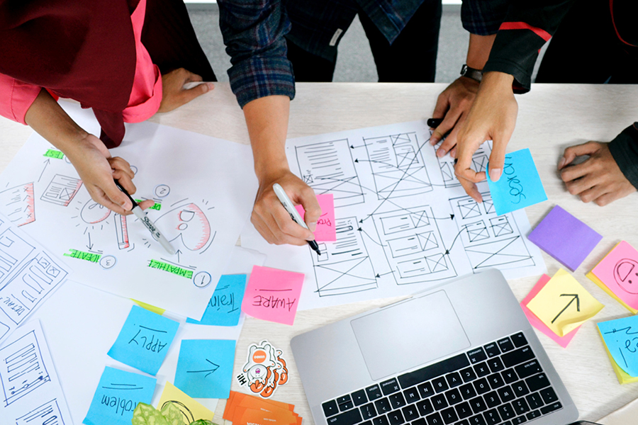 A top-down photo of a diverse team of professionals collaborating around user interface wireframes, paper documents, a computer, sticky notes, and markers on a light-colored wooden surface.