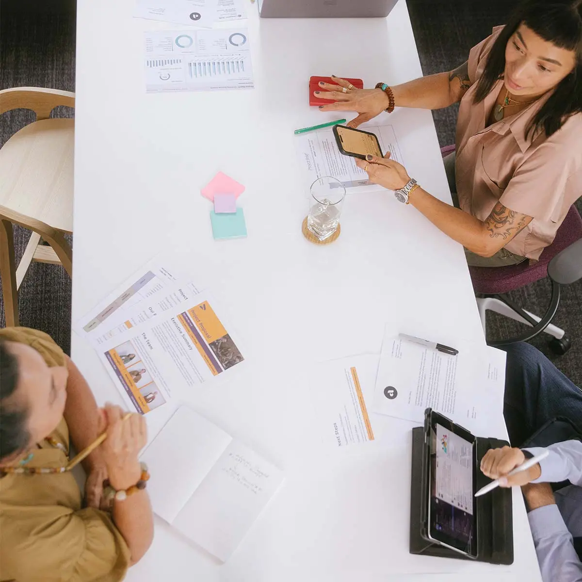 A top-down photo of a diverse team of professionals collaborating around a digital product strategy, including user interface wireframes, paper documents, a computer, sticky notes, and markers on a light-colored wooden surface.