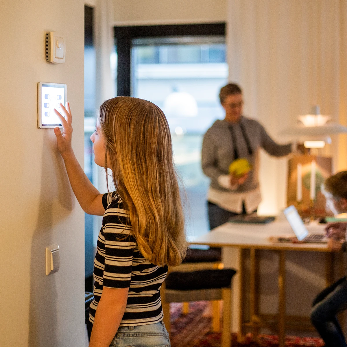 Girl standing in front of a smart thermostat adjusting the settings. Her mother and brother are behind her working at the kitchen table.