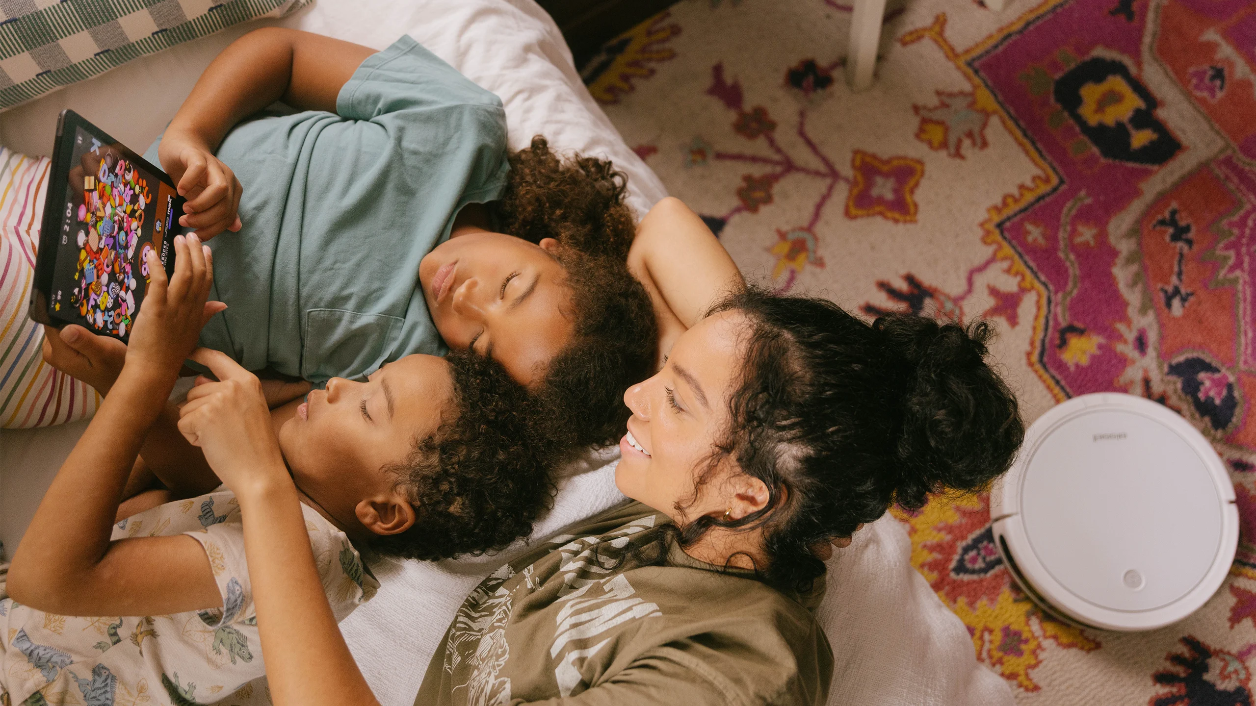 Two sisters and their brother cuddled together on a bed looking at an iPad. Behind them, a robot vacuum is running sweeping the rug in the bedroom.