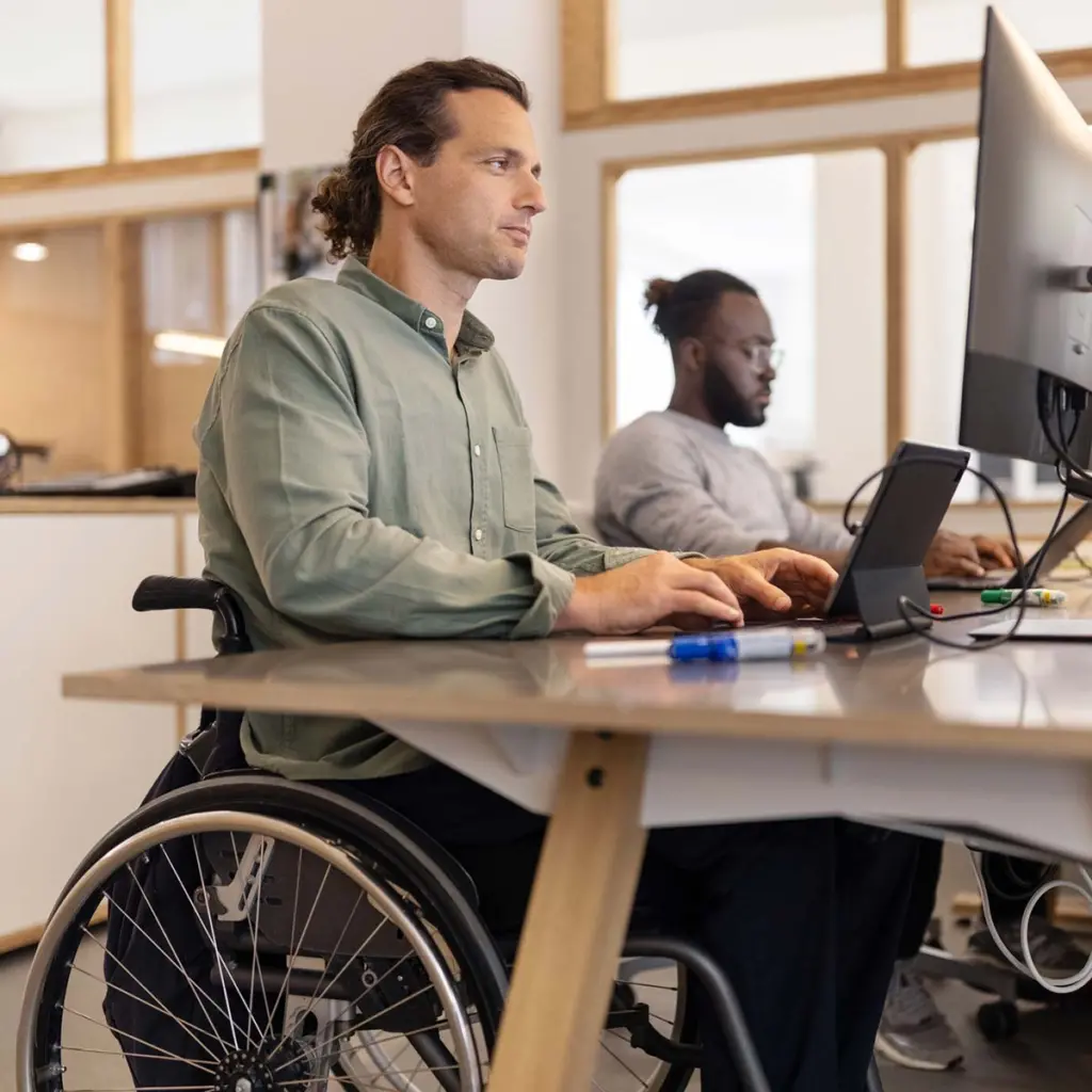 Male executive in wheelchair working on computer in the office with a colleague in background. Multi-ethnic businessmen working at coworking office.