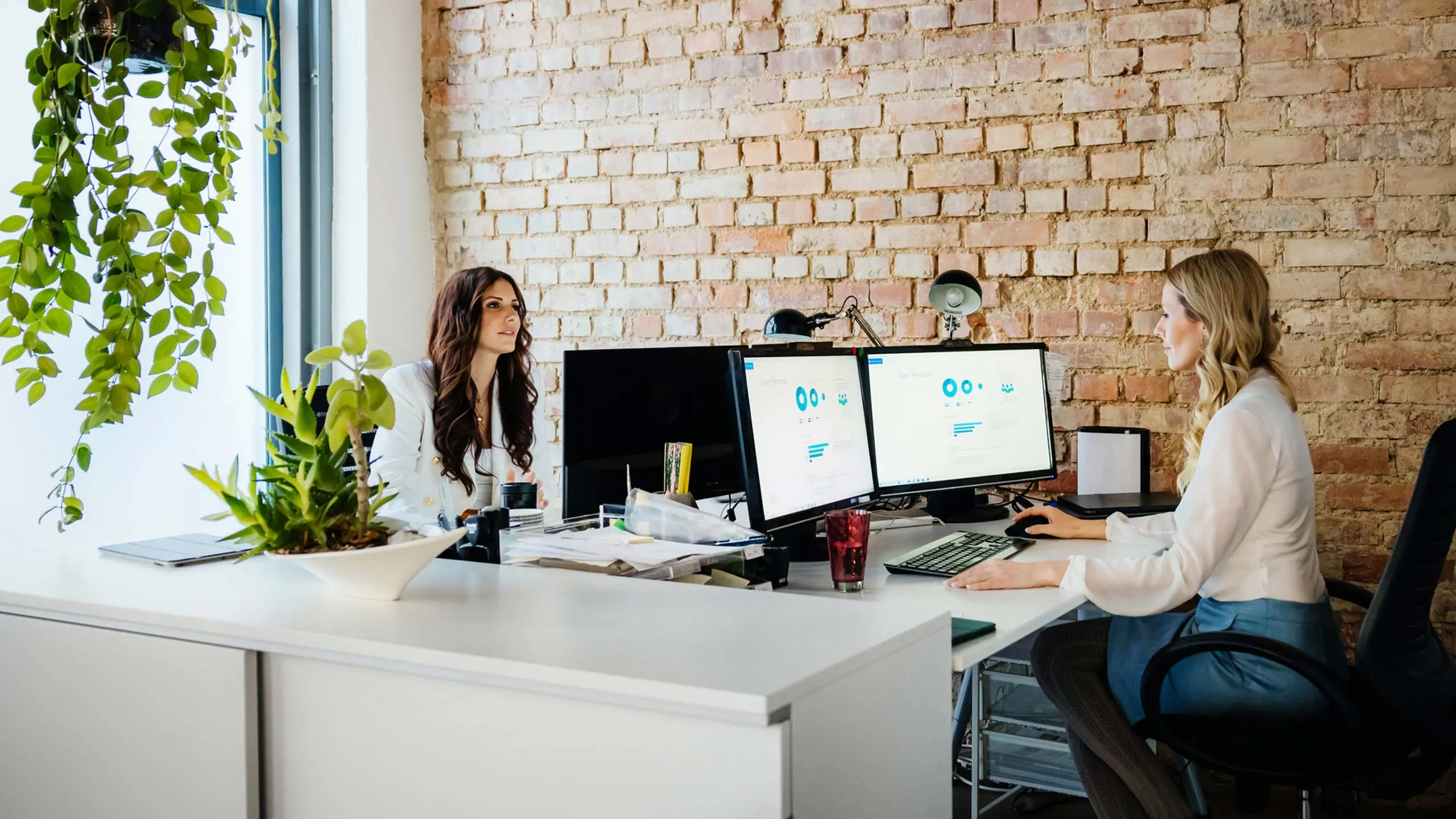 A small, successful, modern business office with two software systems and platforms consulting colleagues sitting at their computer desks, working together. Computer screens showcase data graphs and software development.