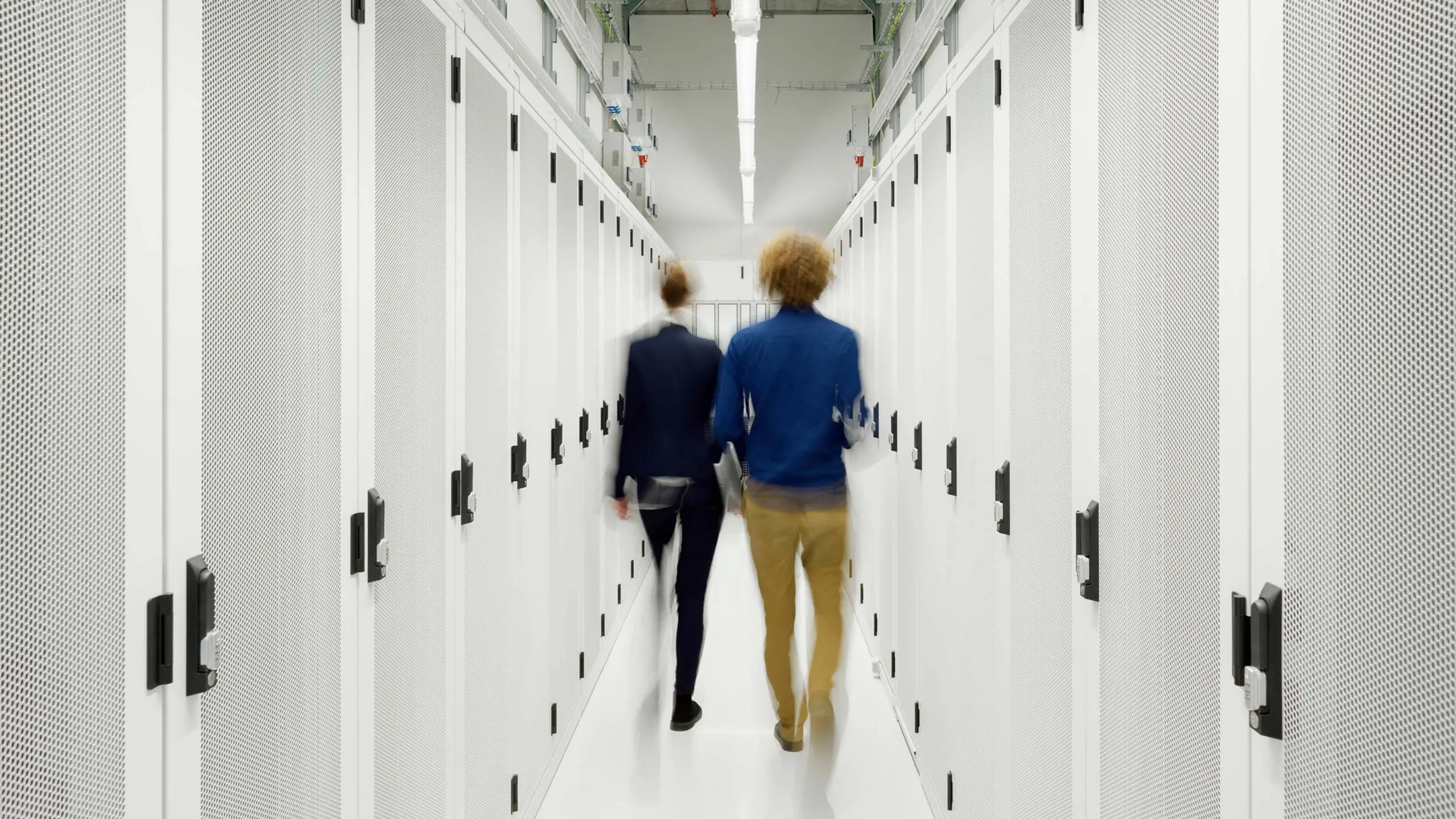 Blurred image of two IT managed services providers walking down bright white data server corridor.