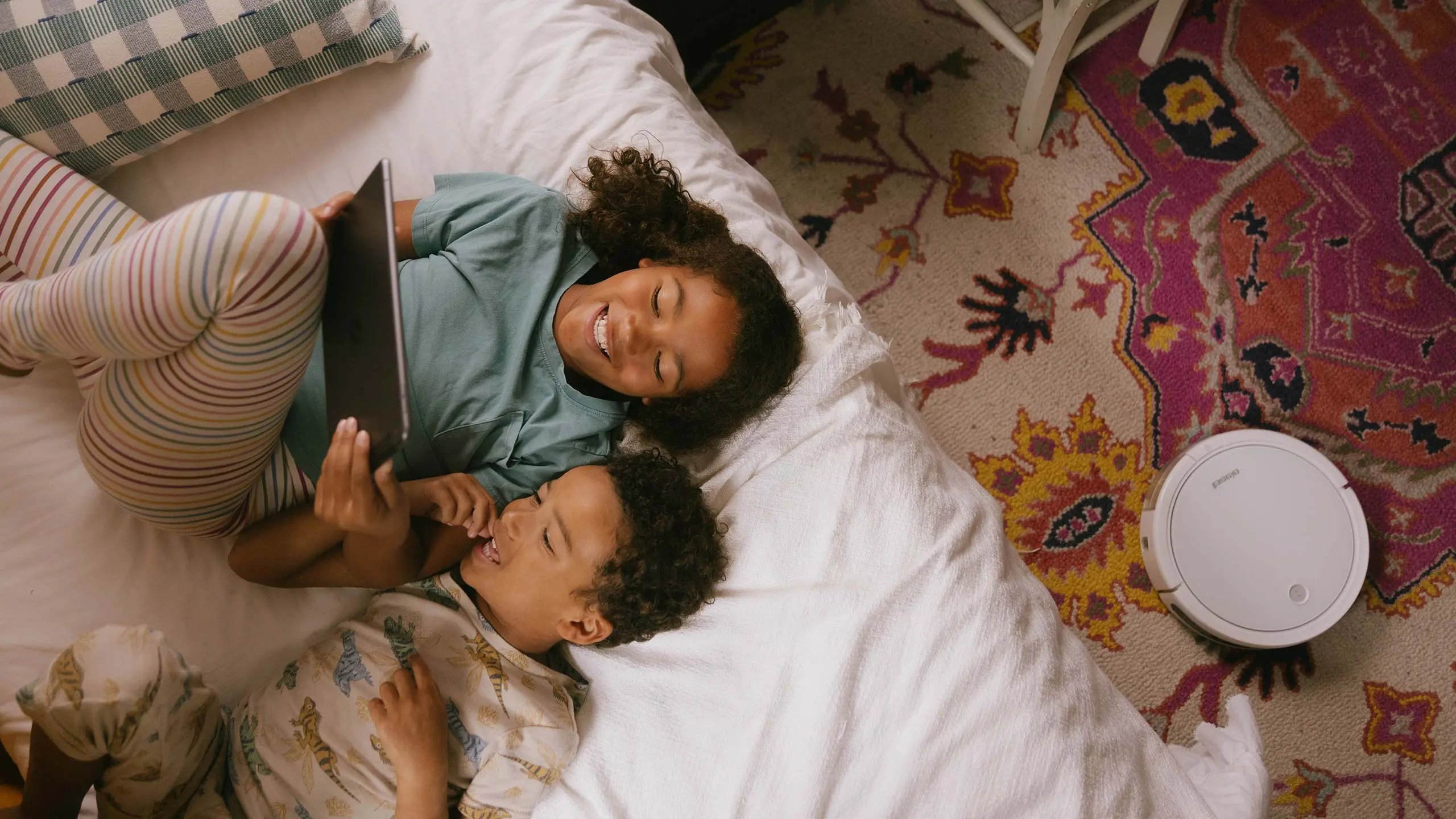 Kids on a bed enjoying technology with a connected IoT product, smart vacuum on the rug behind them.