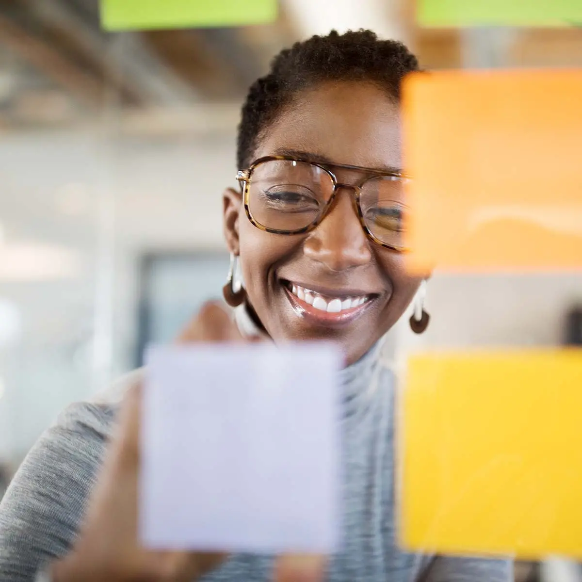 Mid adult female professional writing new ideas on sticky note on glass wall, with a male colleague standing by. Business people seen through glass wall brainstorming ideas in office.