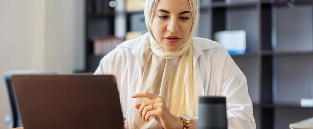 Developer in headscarf using smart speaker while working at her desk in office. Woman using virtual assistant on office table while working on laptop.