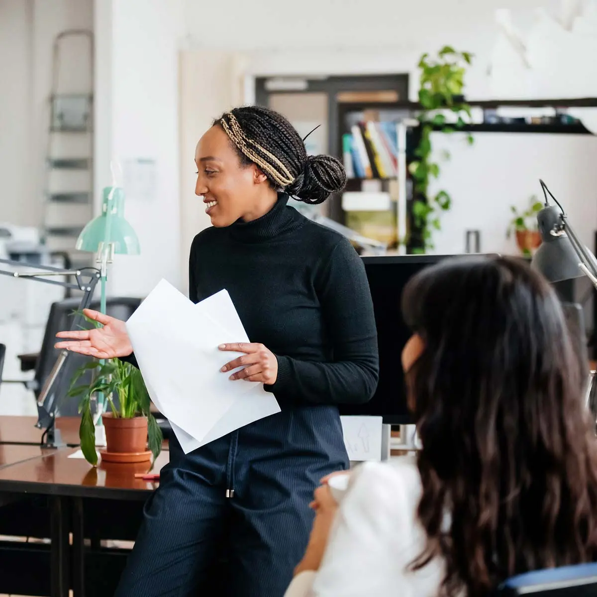 A woman holding some papers and leaning on a computer desk while talking to a group of colleagues during an office meeting.