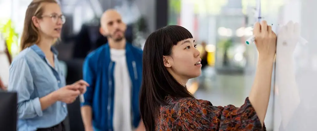 Young businesswoman explaining a business plan to colleague in office. Woman writing on the whiteboard during presentation in hybrid office space.