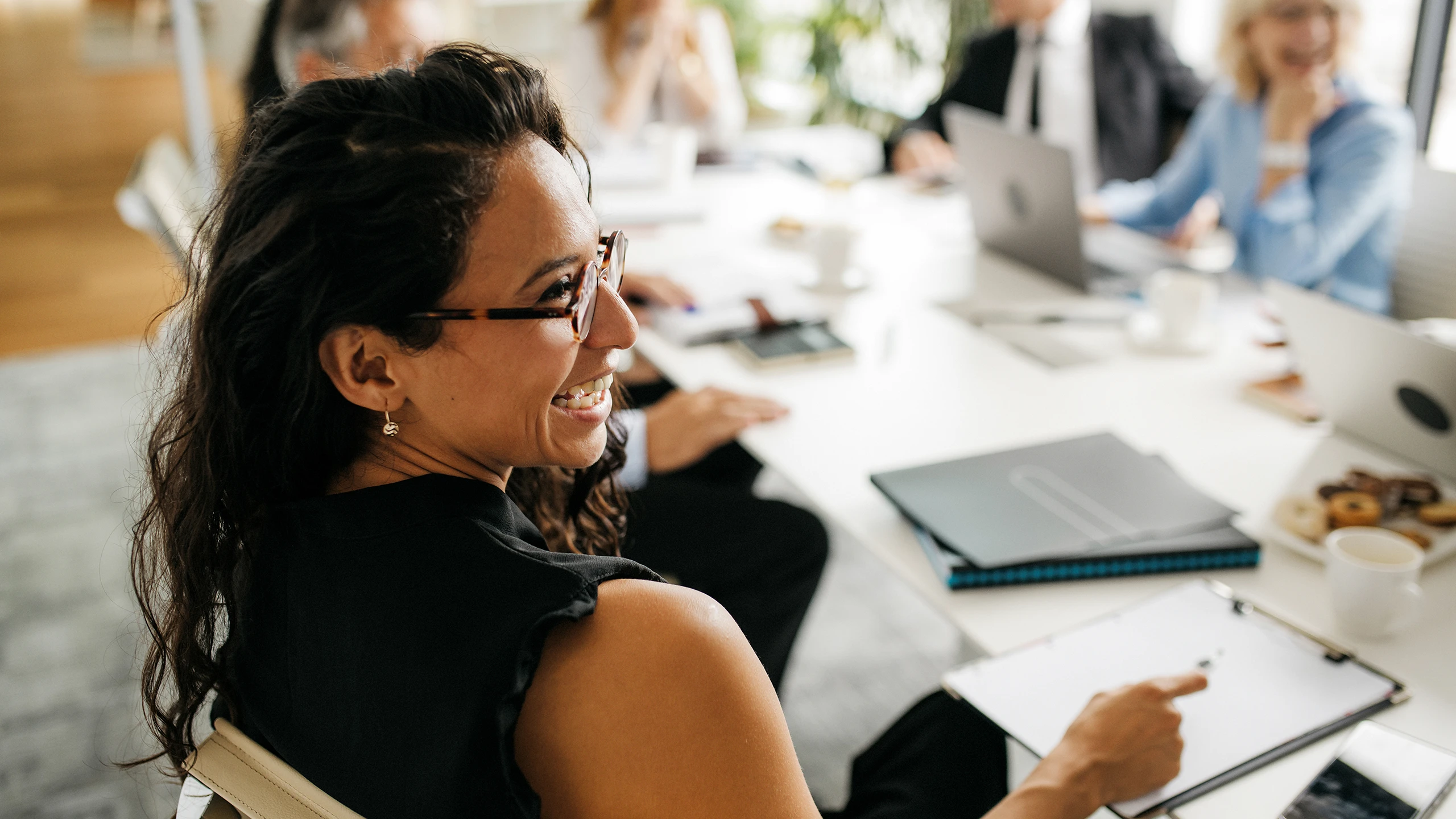 A woman smiling while she sits at a boardroom table in a meeting with a group of people in the background