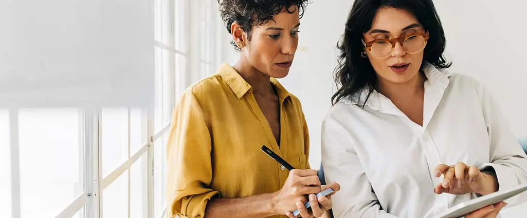 Two business women having a discussion, they're standing in an office and using a tablet. Professional women making a to do list as part of their project planning.