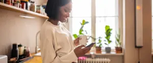 Woman standing in the kitchen holding a cup of coffee and texting on her cell phone. Woman using a mobile phone at home.