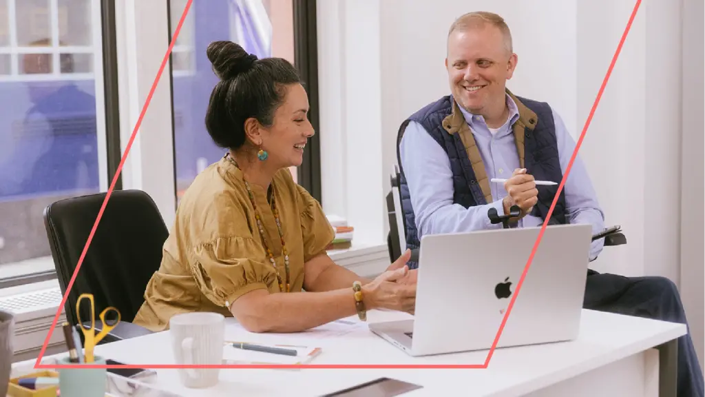 A woman with dark hair in a bun sits at desk smiling down at her laptop on the screen and she discusses something with her colleague. Her colleague is a wheelchair user with light blonde hair. He is holding an Apple pencil and tablet taking notes as he smiles.