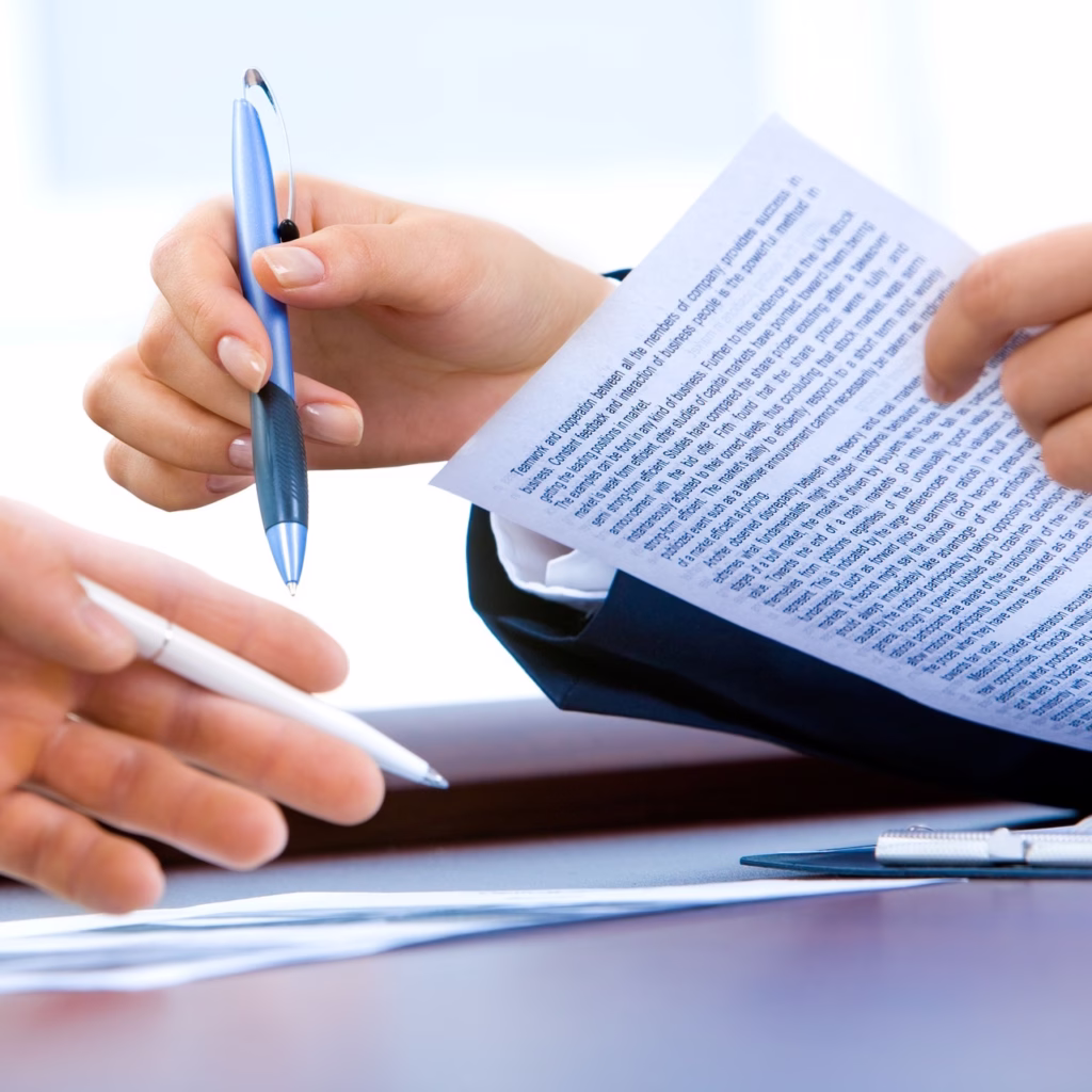 Two hands shown in image as two individuals have a conversation. One, a female, holding a piece of paper with context on the meeting, another a male, carrying a pen.