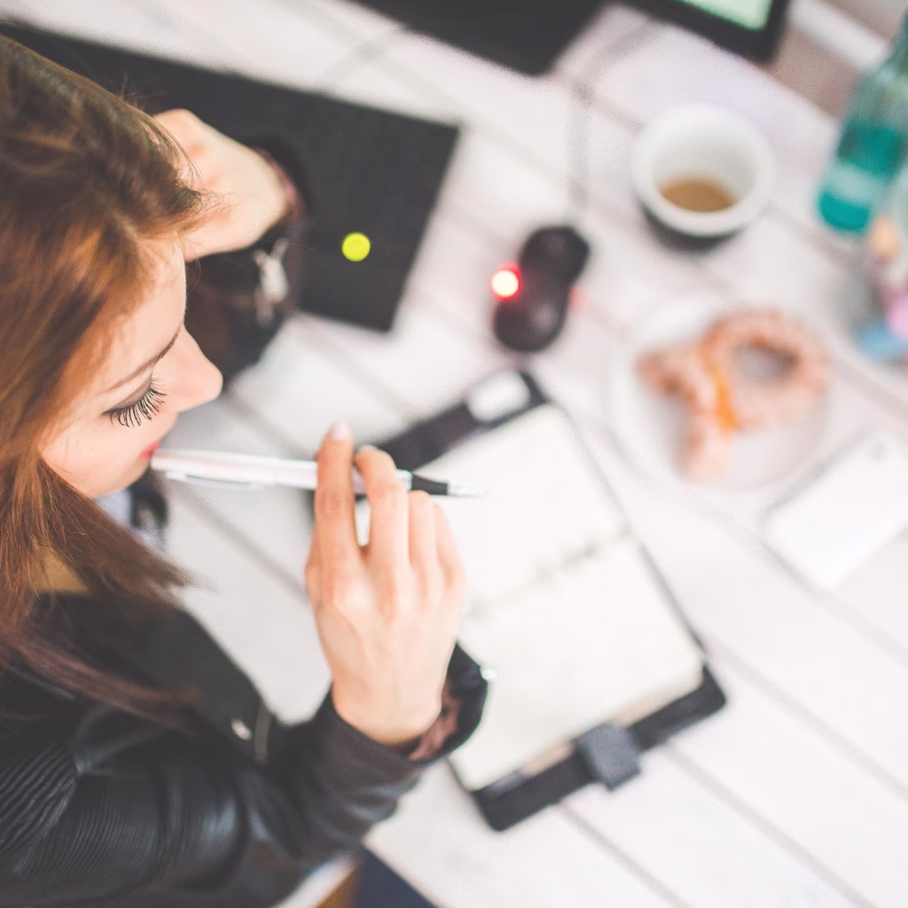 A female looks over her desk while working, pondering. In frame on her desk is her laptop, some food, and her notebook